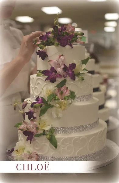 A baker in a white uniform decorating a white three-tiered wedding cake with purple and white alstroemeria flowers in a professional kitchen.