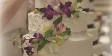 A baker in a white uniform decorating a white three-tiered wedding cake with purple and white alstroemeria flowers in a professional kitchen.