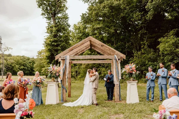 A bride and groom share a kiss under a wooden pergola during an outdoor wedding ceremony, surrounded by bridesmaids, groomsmen, and floral arrangements.