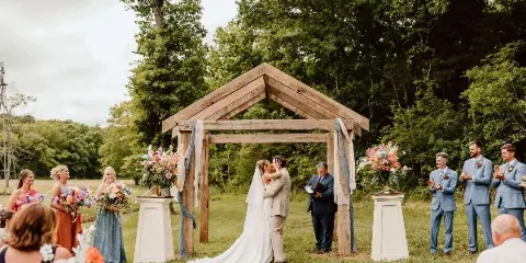 A bride and groom share a kiss under a wooden pergola during an outdoor wedding ceremony, surrounded by bridesmaids, groomsmen, and floral arrangements.