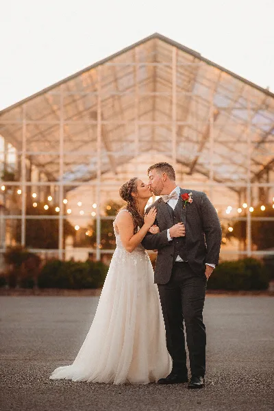 A bride and groom kissing in front of a glass greenhouse structure at sunset.