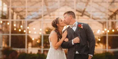 A bride and groom kissing in front of a glass greenhouse structure at sunset.