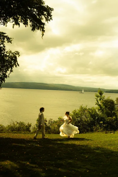 A bride in a white wedding dress and a groom in a light suit walking on a grassy hill overlooking a large body of water with a sailboat in the distance.