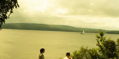 A bride in a white wedding dress and a groom in a light suit walking on a grassy hill overlooking a large body of water with a sailboat in the distance.
