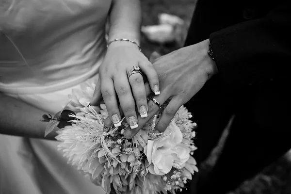A black and white close-up of a bride and groom holding hands, showing their wedding rings and a floral bouquet.