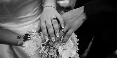 A black and white close-up of a bride and groom holding hands, showing their wedding rings and a floral bouquet.