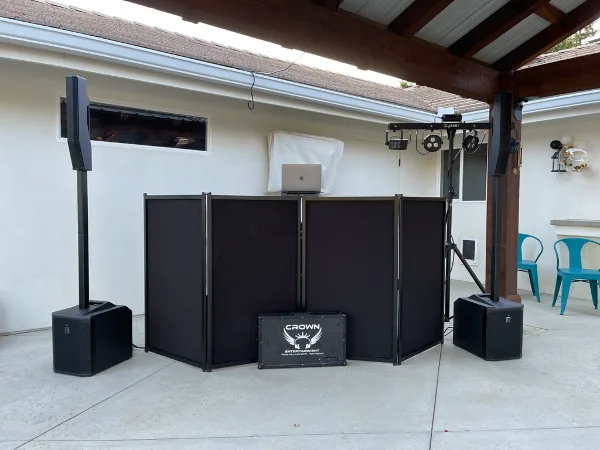 Outdoor DJ setup with black booth panels, tall speakers, lighting truss, and a Crown Entertainment branded flight case.