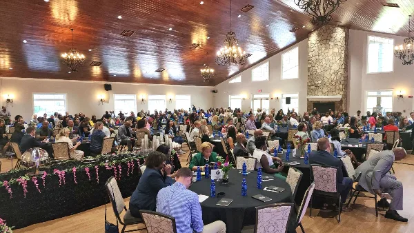 Interior view of a large event hall with round tables, chandeliers, and a stone fireplace, filled with attendees.