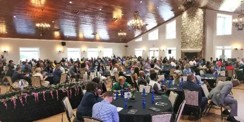 Interior view of a large event hall with round tables, chandeliers, and a stone fireplace, filled with attendees.