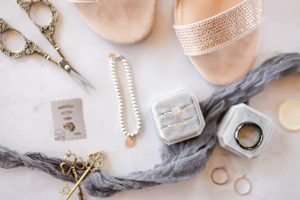 A flat lay photograph of wedding accessories including a diamond ring, wedding bands, pearl necklace, and bridal heels.