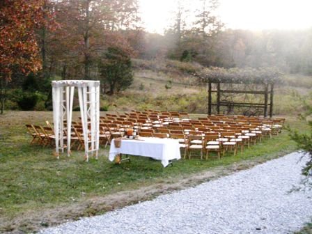Outdoor wedding ceremony setup with a white arch, rows of wooden chairs, and a head table on a grassy field.