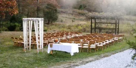 Outdoor wedding ceremony setup with a white arch, rows of wooden chairs, and a head table on a grassy field.