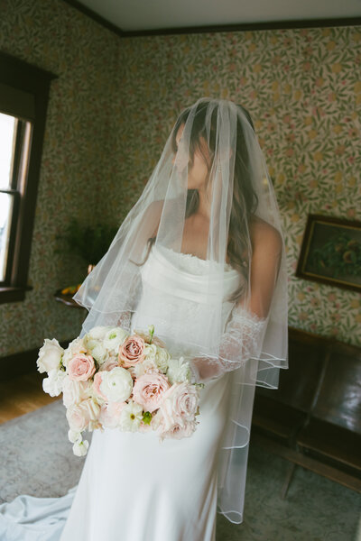 Bride in a white wedding dress and veil holding a bouquet of pink and white flowers indoors.