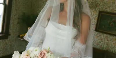 Bride in a white wedding dress and veil holding a bouquet of pink and white flowers indoors.
