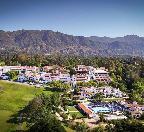 Aerial view of the Ojai Valley Inn resort complex featuring white buildings with red roofs, a swimming pool, and surrounding green grounds and mountains.