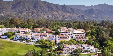 Aerial view of the Ojai Valley Inn resort complex featuring white buildings with red roofs, a swimming pool, and surrounding green grounds and mountains.