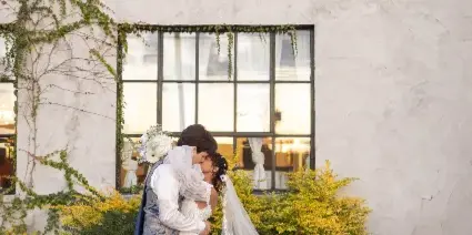 A bride and groom sharing a kiss outdoors in front of a white textured wall with greenery.