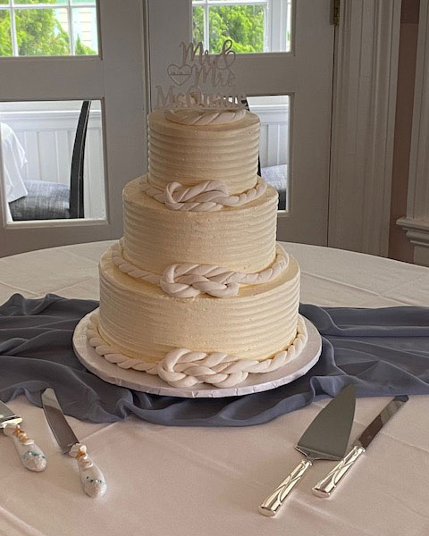A three-tiered white wedding cake with rope-style frosting details and a topper, displayed on a table with cake serving tools.