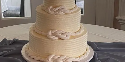 A three-tiered white wedding cake with rope-style frosting details and a topper, displayed on a table with cake serving tools.