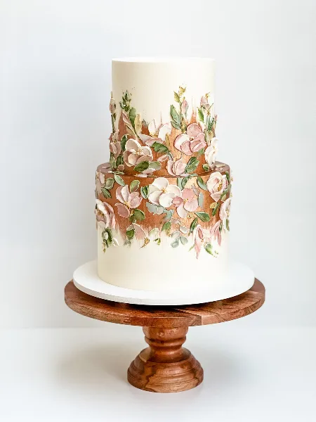 A two-tier white wedding cake decorated with hand-piped pink and white buttercream flowers and green leaves, displayed on a wooden pedestal stand.
