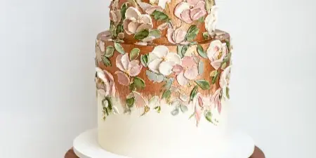 A two-tier white wedding cake decorated with hand-piped pink and white buttercream flowers and green leaves, displayed on a wooden pedestal stand.