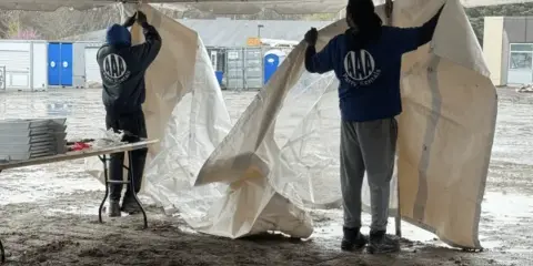 Two workers in branded shirts setting up a white event tent and handling linens outdoors.