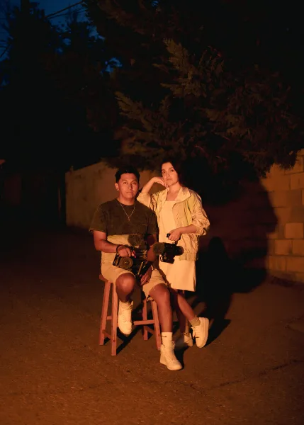 A man and a woman sitting on stools outdoors at night, holding professional video cameras.