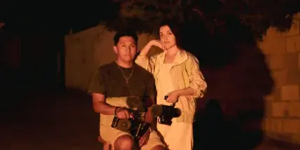A man and a woman sitting on stools outdoors at night, holding professional video cameras.