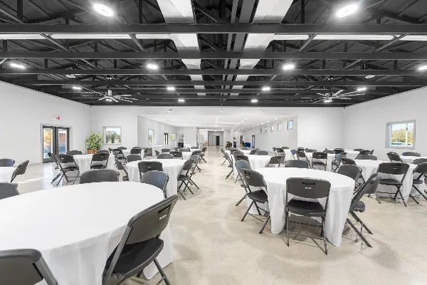 Interior view of a modern event venue with round tables, white tablecloths, black chairs, and an exposed dark ceiling with lighting and fans.