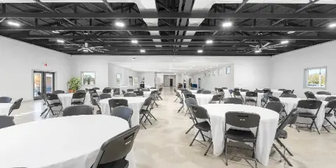 Interior view of a modern event venue with round tables, white tablecloths, black chairs, and an exposed dark ceiling with lighting and fans.