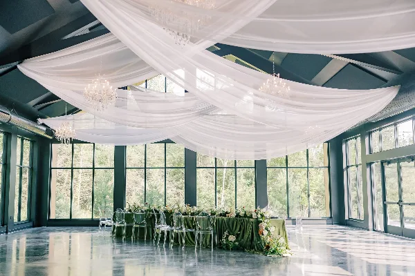 A wide shot of a wedding venue featuring elaborate white ceiling draping, hanging crystal chandeliers, and a decorated head table.