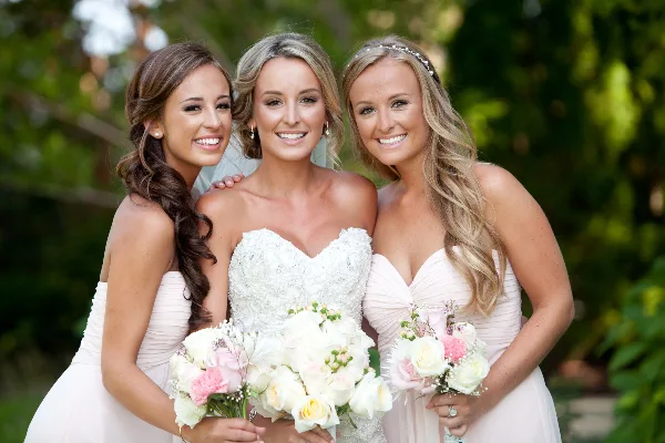 A bride and two bridesmaids smiling with styled hair and makeup, holding bouquets.