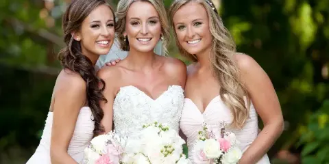 A bride and two bridesmaids smiling with styled hair and makeup, holding bouquets.