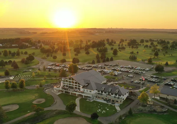 Aerial view of a large white venue building with a grey roof, surrounded by a green golf course and a parking lot at sunset.