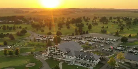 Aerial view of a large white venue building with a grey roof, surrounded by a green golf course and a parking lot at sunset.