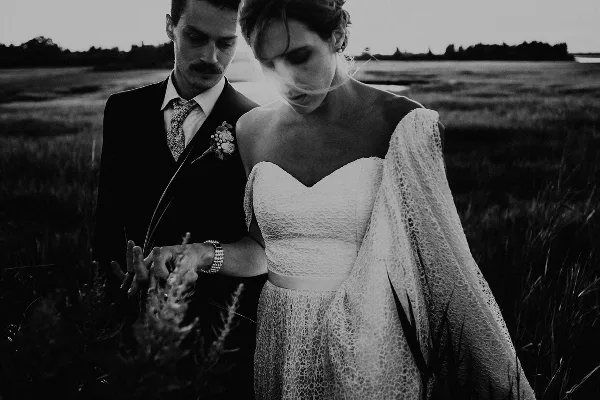Black and white photograph of a bride and groom standing in a grassy field, with the groom placing a ring on the bride's finger.