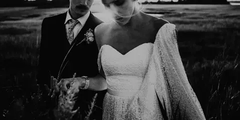 Black and white photograph of a bride and groom standing in a grassy field, with the groom placing a ring on the bride's finger.