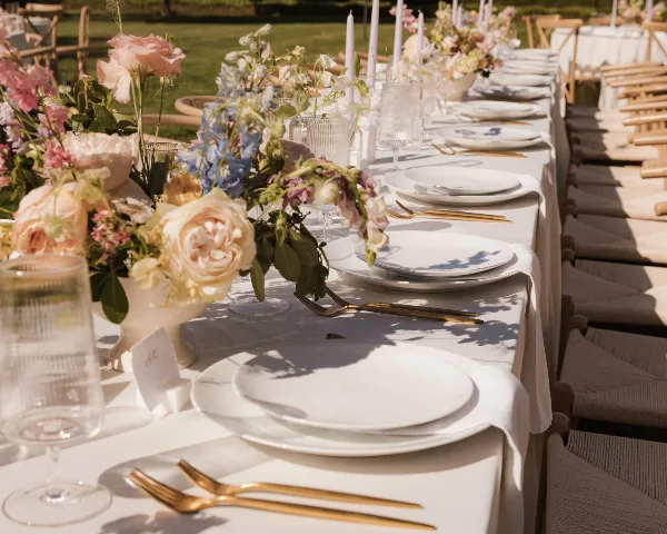 A long outdoor event table set with white linens, gold flatware, glassware, and floral centerpieces.