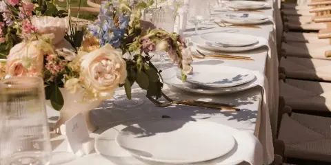 A long outdoor event table set with white linens, gold flatware, glassware, and floral centerpieces.
