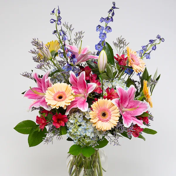 A vibrant floral arrangement featuring pink lilies, gerbera daisies, delphiniums, and hydrangeas in a clear glass vase.