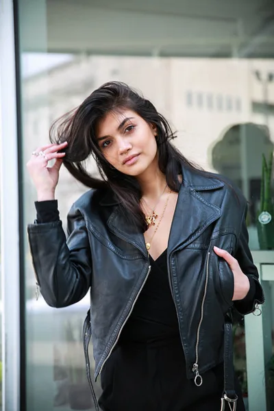 A professional fashion portrait of a woman wearing a black leather jacket and gold jewelry, posing outdoors.