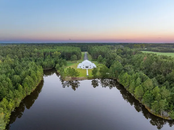 Aerial view of a white wedding venue building on a lawn next to a lake, surrounded by pine trees at sunset.