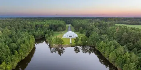 Aerial view of a white wedding venue building on a lawn next to a lake, surrounded by pine trees at sunset.