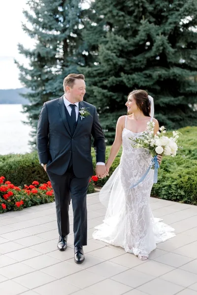 A bride and groom holding hands and walking on a stone patio near a lake with evergreen trees in the background.