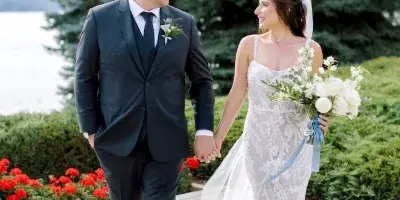 A bride and groom holding hands and walking on a stone patio near a lake with evergreen trees in the background.