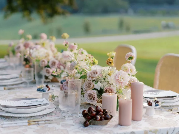 A wedding table setting featuring a romantic floral centerpiece with pink and white blooms, candles, and glassware on a patterned tablecloth.