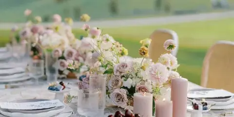 A wedding table setting featuring a romantic floral centerpiece with pink and white blooms, candles, and glassware on a patterned tablecloth.