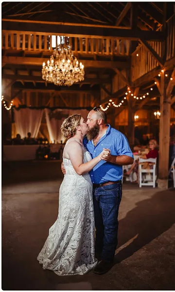 A bride and groom sharing a first dance in a rustic barn venue illuminated by a large chandelier and string lights.