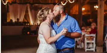 A bride and groom sharing a first dance in a rustic barn venue illuminated by a large chandelier and string lights.