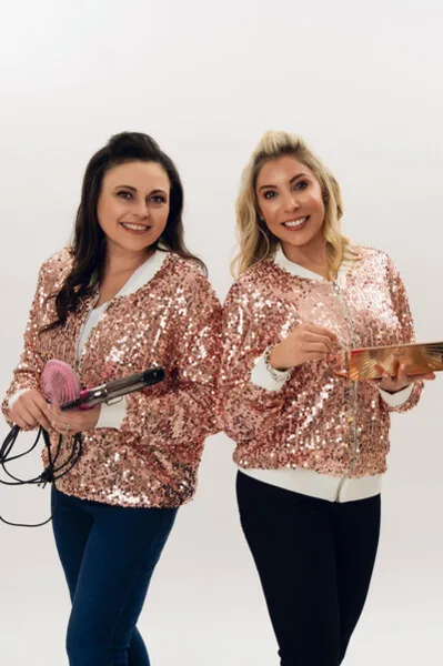 Two women in matching sequined jackets holding a hair straightener and a round brush.